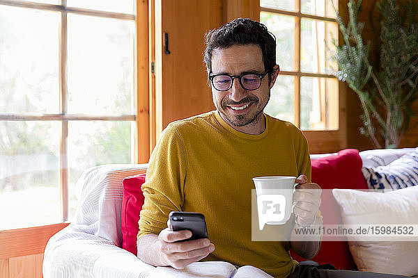 Smiling man holding coffee using smart phone while sitting on sofa in log cabin