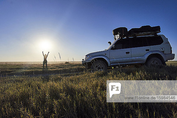 Frau steht mit erhobenen Armen neben einem Geländewagen in den Makgadikgadi Pans  Botsuana