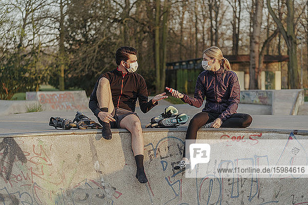 Frau gibt Mann Handdesinfektionsmittel  während er in einem Skateboard-Park während COVID-19 sitzt