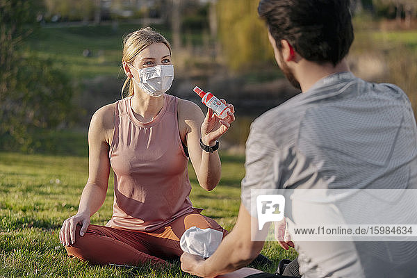 Frau mit Gesichtsmaske zeigt Mann Handdesinfektionsmittel  während er im Park sitzt
