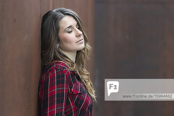 Portrait of young woman wearing red plaid shirt leaning against brown wall