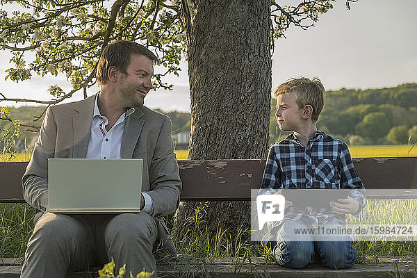 Smiling man looking at son sitting with laptop and digital tablet on bench