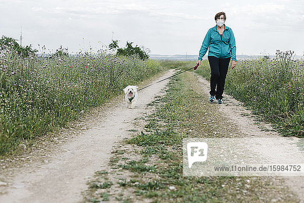 Ältere Frau mit Schutzmaske geht mit ihrem Hund auf einem Feldweg spazieren