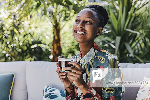 Portrait of happy young woman sitting on couch in garden drinking black coffee