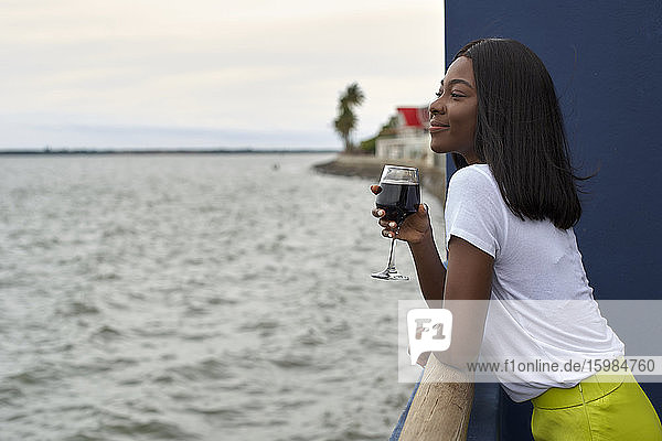 Glückliche junge Frau mit einem Glas Rotwein und Blick auf das Meer