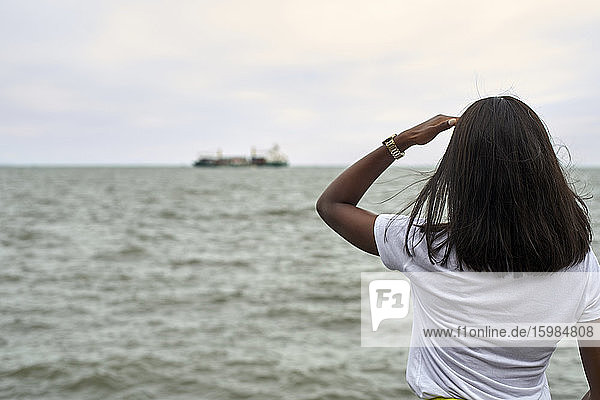 Rückenansicht einer jungen Frau vor dem Meer mit Blick auf ein Schiff am Horizont