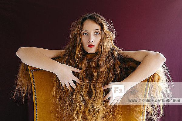 Porträt einer selbstbewussten jungen Frau mit langen braunen gewellten Haaren  die sich auf einen Stuhl vor einem farbigen Hintergrund stützt