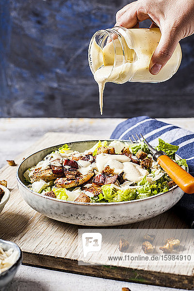Hand einer Person  die das Dressing über eine Schüssel mit Caesar-Salat mit Römersalat  Parmesankäse  Speck  Hähnchenbrust und Croutons gießt