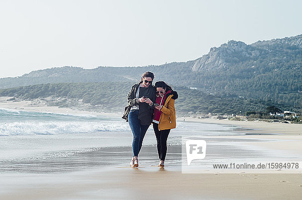 Volle Länge von glücklichen Freundinnen  die Mobiltelefone benutzen  während sie am Strand in Tarifa  Spanien  spazieren gehen