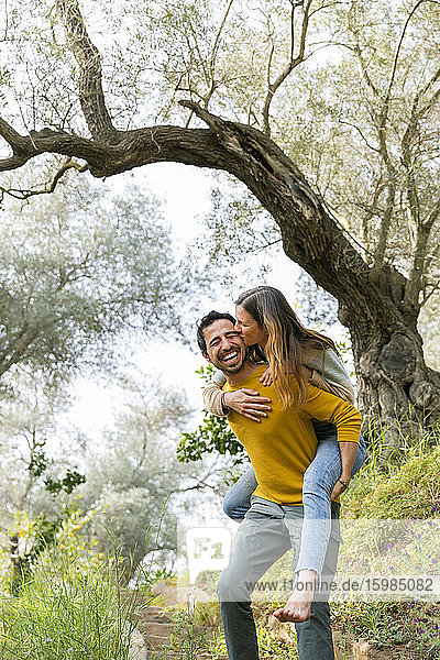 Fröhlicher Mann  der seine romantische Freundin huckepack nimmt  während er auf dem Lande gegen Bäume steht