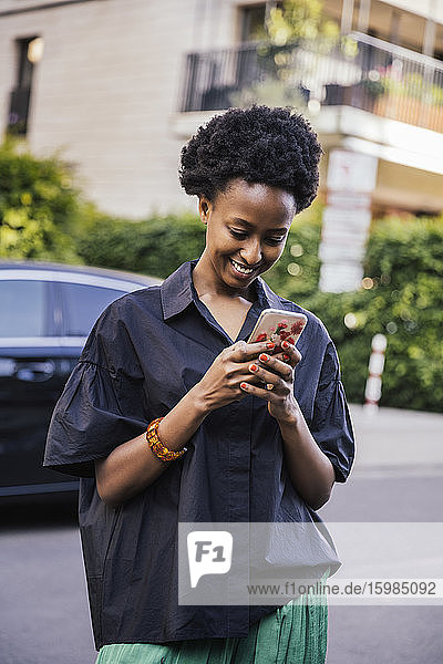 Porträt einer lächelnden jungen Frau  die auf der Straße steht und auf ihr Mobiltelefon schaut