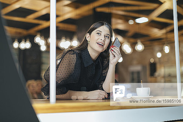 Lächelnde schöne junge Frau  die ihr Smartphone hält und in ein beleuchtetes Café schaut