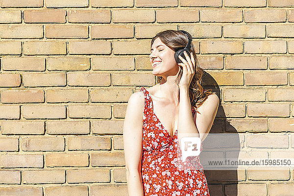 Happy young woman listening to music and leaning on brick wall