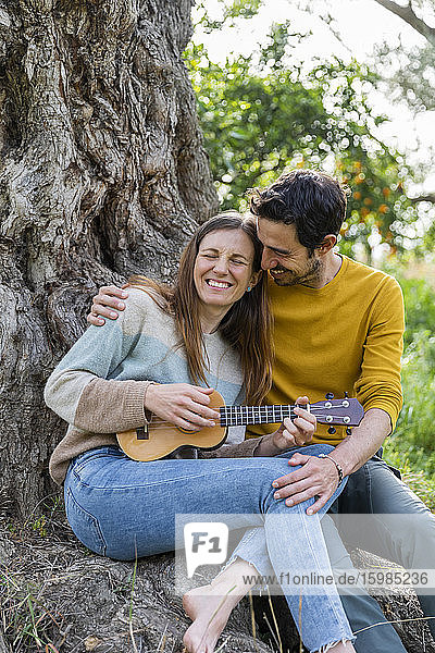 Loving man embracing happy girlfriend playing guitar while sitting against tree trunk