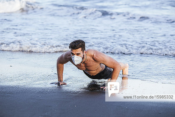 Shirtless mid adult man wearing face mask while doing push-ups at beach during sunset