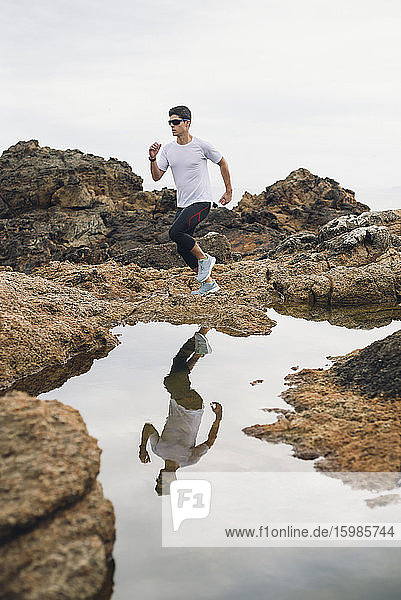 Männlicher Trailrunner in voller Länge  der auf Felsen läuft  mit seinem Spiegelbild über dem Wasser an der Küste  Ferrol  Galicien  Spanien