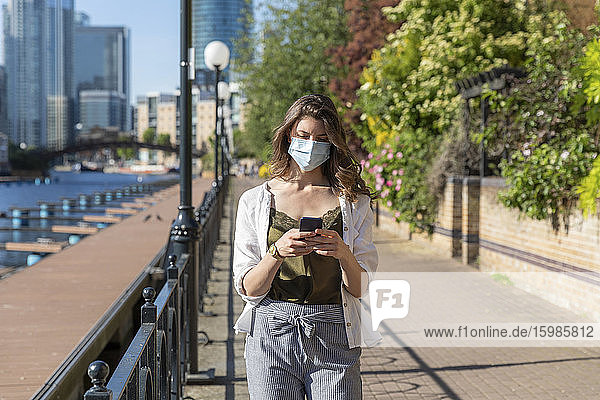 Junge Frau mit Schutzmaske  die spazieren geht und ein Smartphone benutzt