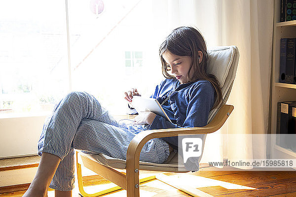 Junges Mädchen sieht sich ein Online-Video auf einem digitalen Tablet an  während sie zu Hause am Fenster sitzt
