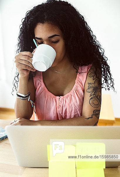 Junge Frau arbeitet von zu Hause aus mit Laptop und trinkt Kaffee