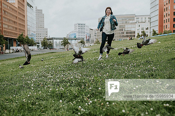 Deutschland  Berlin  Junge Frau läuft auf Gras und überrascht Taubenschwarm