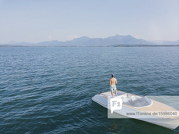Germany  Bavaria  Shirtless man standing on motorboat floating in Chiemsee lake admiring view of distant Chiemgau Alps