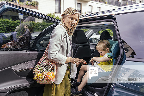 Porträt einer lächelnden Frau  die vor einem Auto wartet  während ihr kleiner Sohn die Sitzglocke des Kindersitzes befestigt