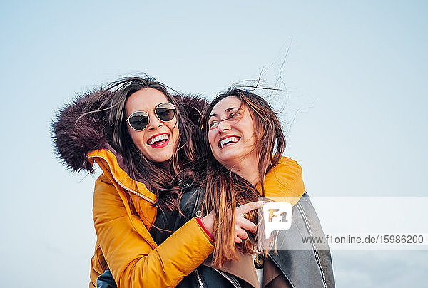 Fröhliche Frau  die ihre Freundin huckepack nimmt  während sie am Strand gegen den Himmel in Tarifa  Spanien  Spaß hat