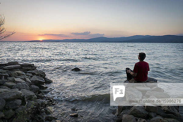 Rear view of mature man looking at Lake Trasimeno while sitting on rocks at sunset  Isola Maggiore  Italy