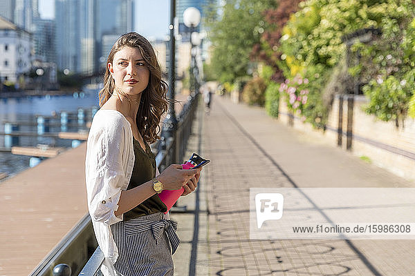 Junge Frau lehnt sich mit ihrem Smartphone an das Geländer der Promenade