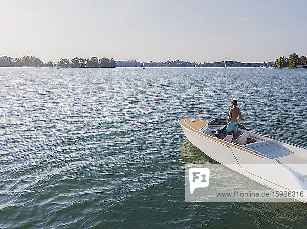 Germany  Bavaria  Shirtless man standing on motorboat floating in Chiemsee lake admiring distant landscape