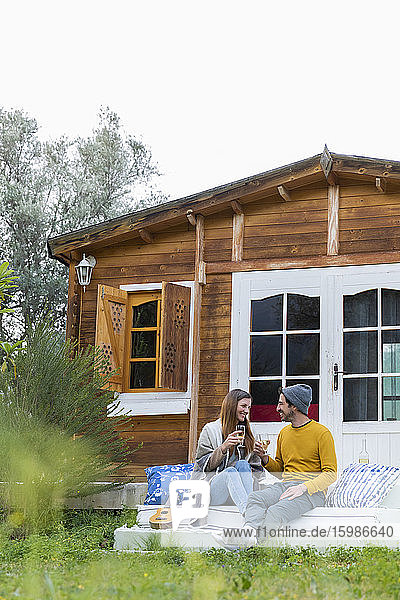 Glückliches Paar mit Weingläsern in der Hand  das auf den Stufen einer Blockhütte sitzt