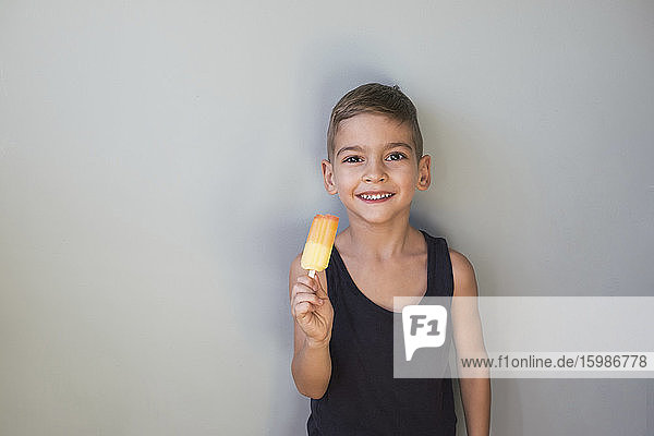 Porträt eines niedlichen lächelnden Jungen  der ein Eis am Stiel isst und an der Wand steht