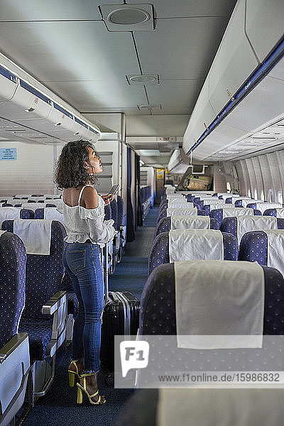 Junger weiblicher Passagier auf der Suche nach einem Sitzplatz in voller Länge  während er im Gang eines Flugzeugs steht
