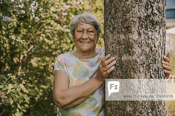 Lächelnde aktive ältere Frau  die an einem Baum vor Pflanzen im Garten steht