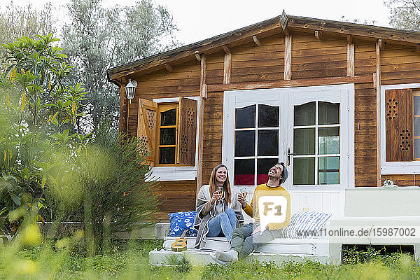 Fröhliches Paar mit Weingläsern in der Hand  das lachend auf den Stufen einer Blockhütte sitzt