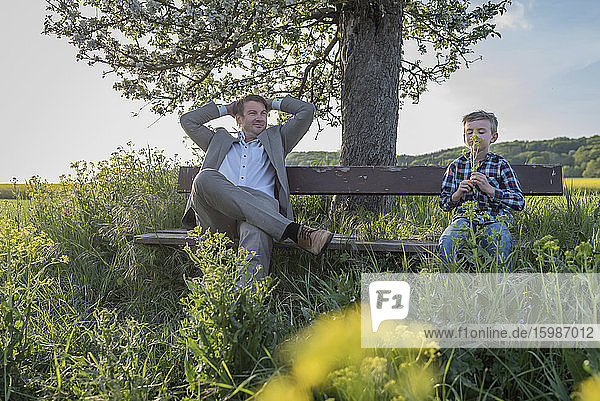 Relaxed man looking away while sitting by son smelling flowers on bench at park against sky