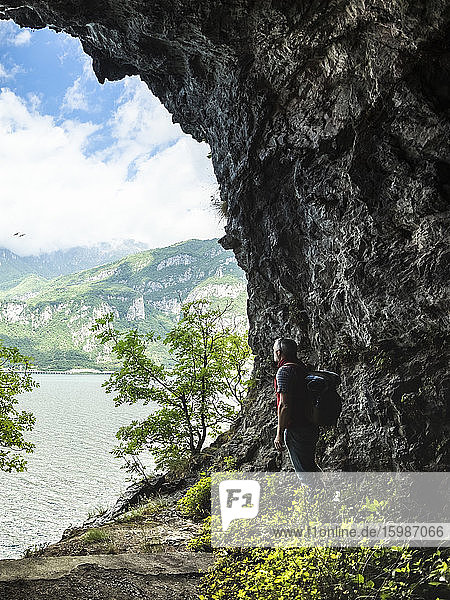Mature man with backpack standing by rock formation while looking at Lake Como from cave entrance