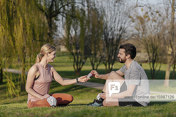 Smiling man giving hand sanitizer to woman while sitting on grass at park