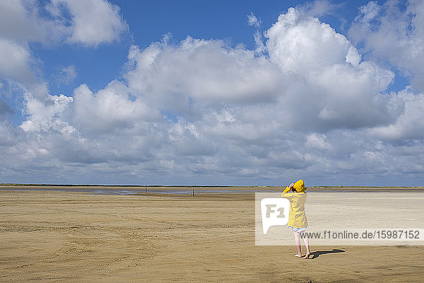 Full length rear view of teenage girl wearing yellow raincoat while walking on sand at beach during sunny day