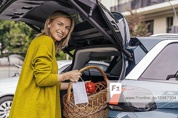 Porträt einer glücklichen Frau mit Schutzmaske  die ihren Einkauf in den Kofferraum ihres Autos legt