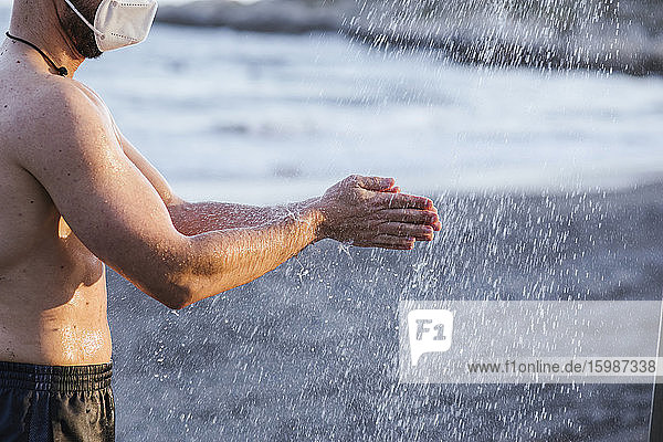Shirtless Mitte erwachsenen Mann mit Gesichtsmaske Waschen der Hände unter der Dusche am Strand bei Sonnenuntergang