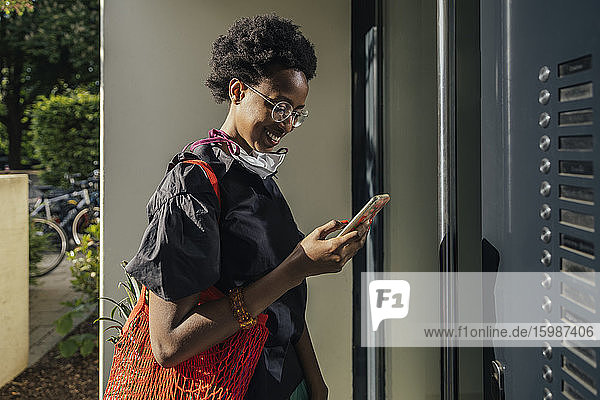 Lächelnde junge Frau mit textiler Schutzmaske steht vor der Eingangstür und schaut auf ihr Smartphone