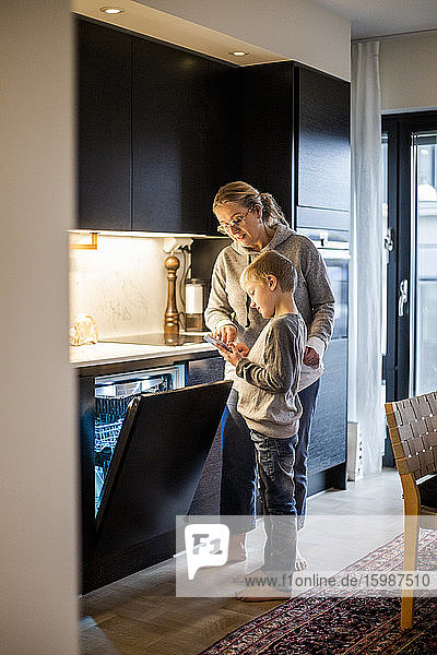 Mutter bringt Sohn bei  eine mobile App zu benutzen  während er zu Hause in der Küche den Geschirrspüler bedient