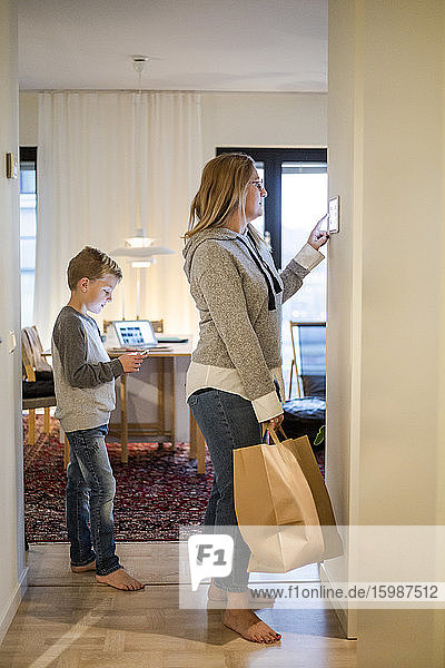 Mutter mit Einkaufstasche mit digitalem Tablett an der Wand  während der Sohn zu Hause steht