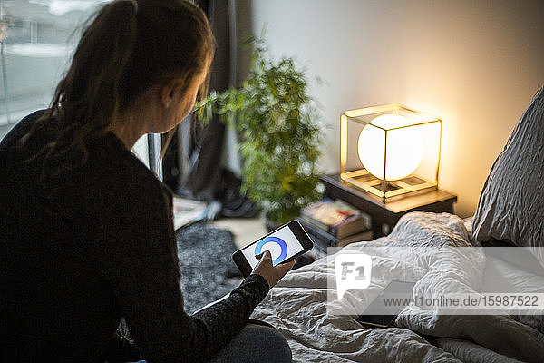 Teenager-Mädchen benutzt Smartphone-App bei der Einstellung der Beleuchtungsanlage im Schlafzimmer in einem Smart Home