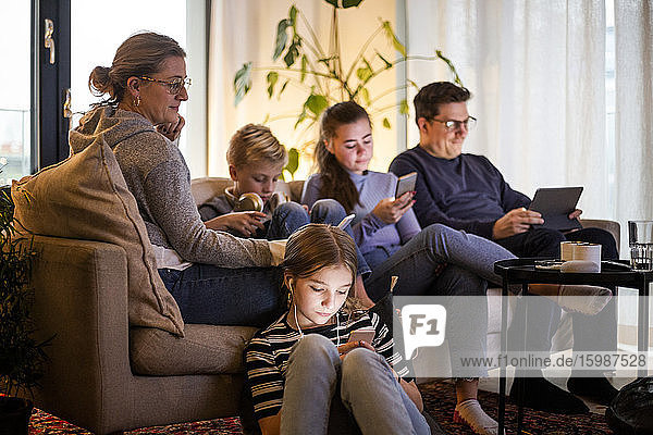 Eltern sitzen mit Kindern zusammen  während sie drahtlose Technologien im Wohnzimmer eines modernen Zuhauses nutzen