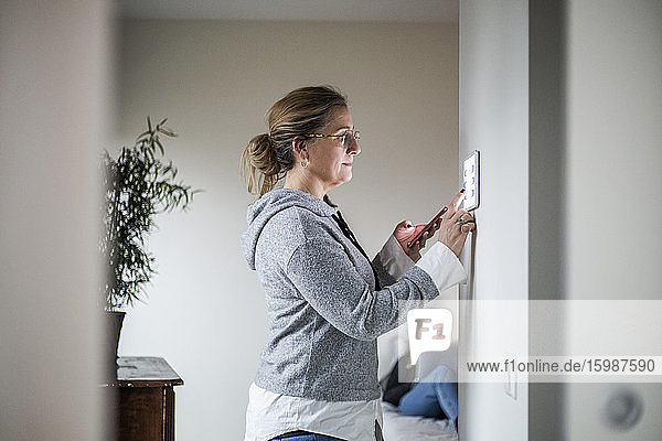 Mutter hält Smartphone in der Hand  während sie ein digitales Tablet an der Wand hält  während die Tochter im Hintergrund im Smart Home ist