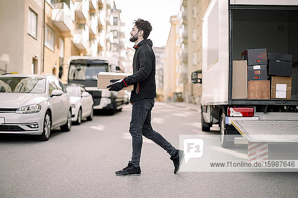 Seitenansicht eines jungen Boten  der eine Kiste trägt  während er in der Stadt auf der Straße geht
