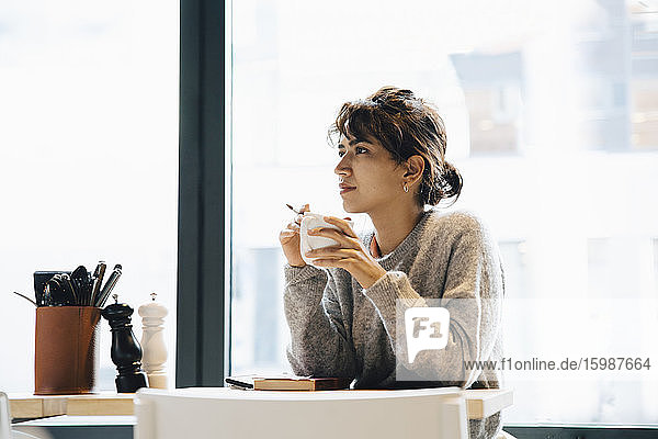 Thoughtful woman looking away while sitting with coffee cup at table in cafeteria