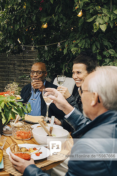 Lächelnde ältere Frau sitzt mit männlichen Freunden zusammen  während sie während der Dinnerparty im Hinterhof am Esstisch anstößt